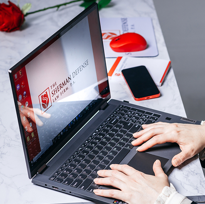 Working on a Laptop with The Sherman Defense Branding A person's hands typing on a laptop whose screen displays the logo and name of "The Sherman Defense Law Firm, P.A." The laptop is on a marble surface next to a red rose and a smartphone.
