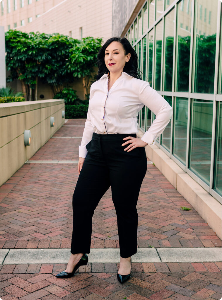 Professional Woman in Business Attire A professional woman with dark hair wearing a white button-down shirt and black trousers, standing on a brick walkway in an urban setting with a glass-fronted building and landscaping behind her.