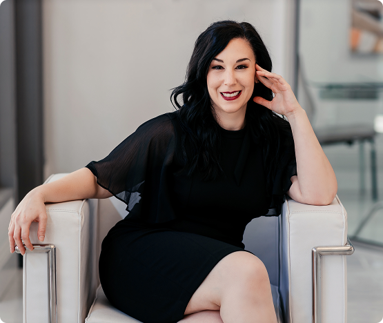 Smiling Professional Woman Portrait A professional woman with dark hair, wearing a black dress with sheer sleeves, is seated in a white modern armchair, smiling directly at the camera.